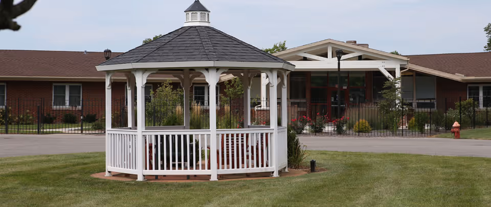 White wooden gazebo on a grassy lawn in front of a single-story brick senior living building entrance.