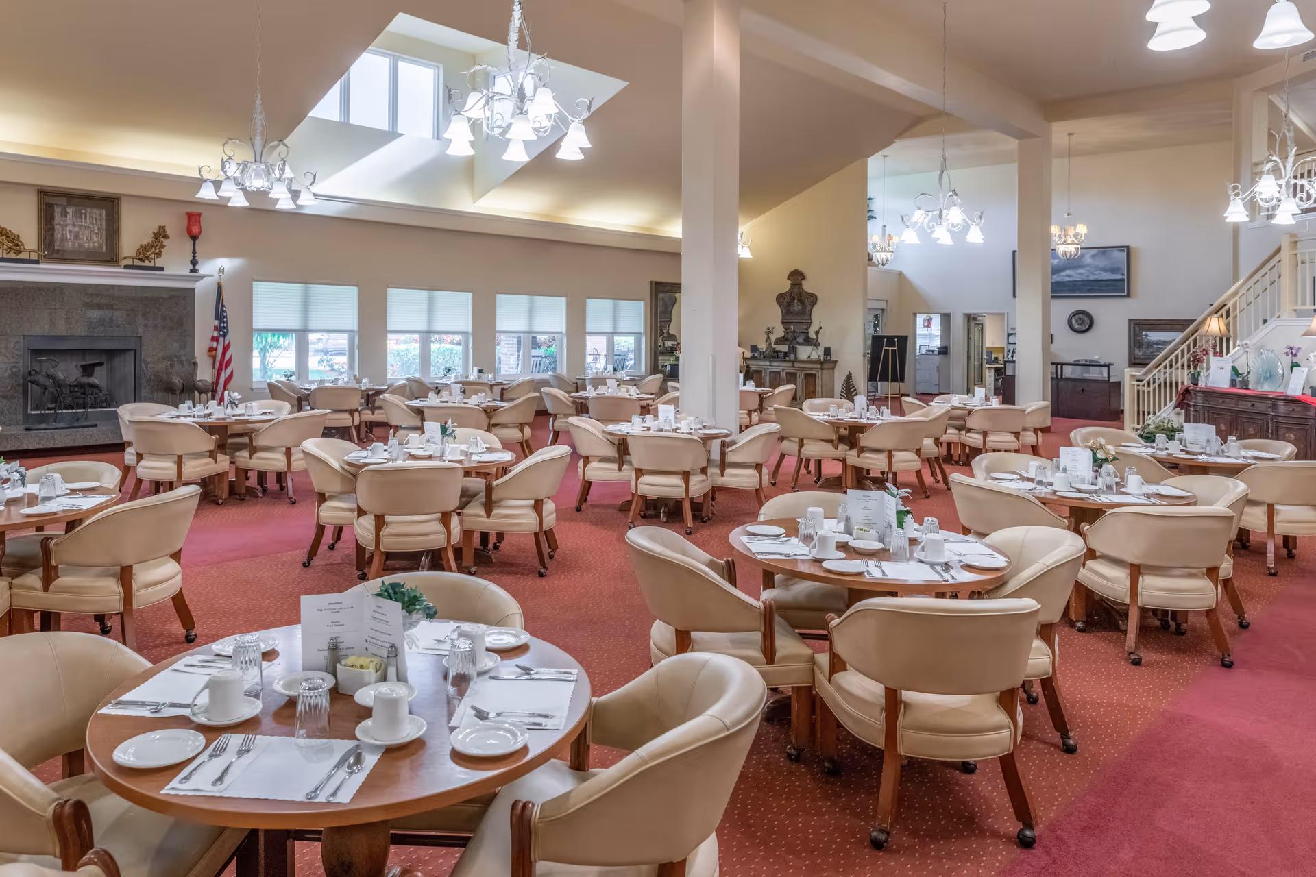 Spacious dining room with multiple round tables set with white cups, plates, and silverware. Beige cushioned chairs surround each table. The room features a red carpet, large windows with blinds, chandeliers hanging from the ceiling, and a fireplace with an American flag beside it. There are decorative items and framed pictures on the walls, and a staircase is visible in the background.