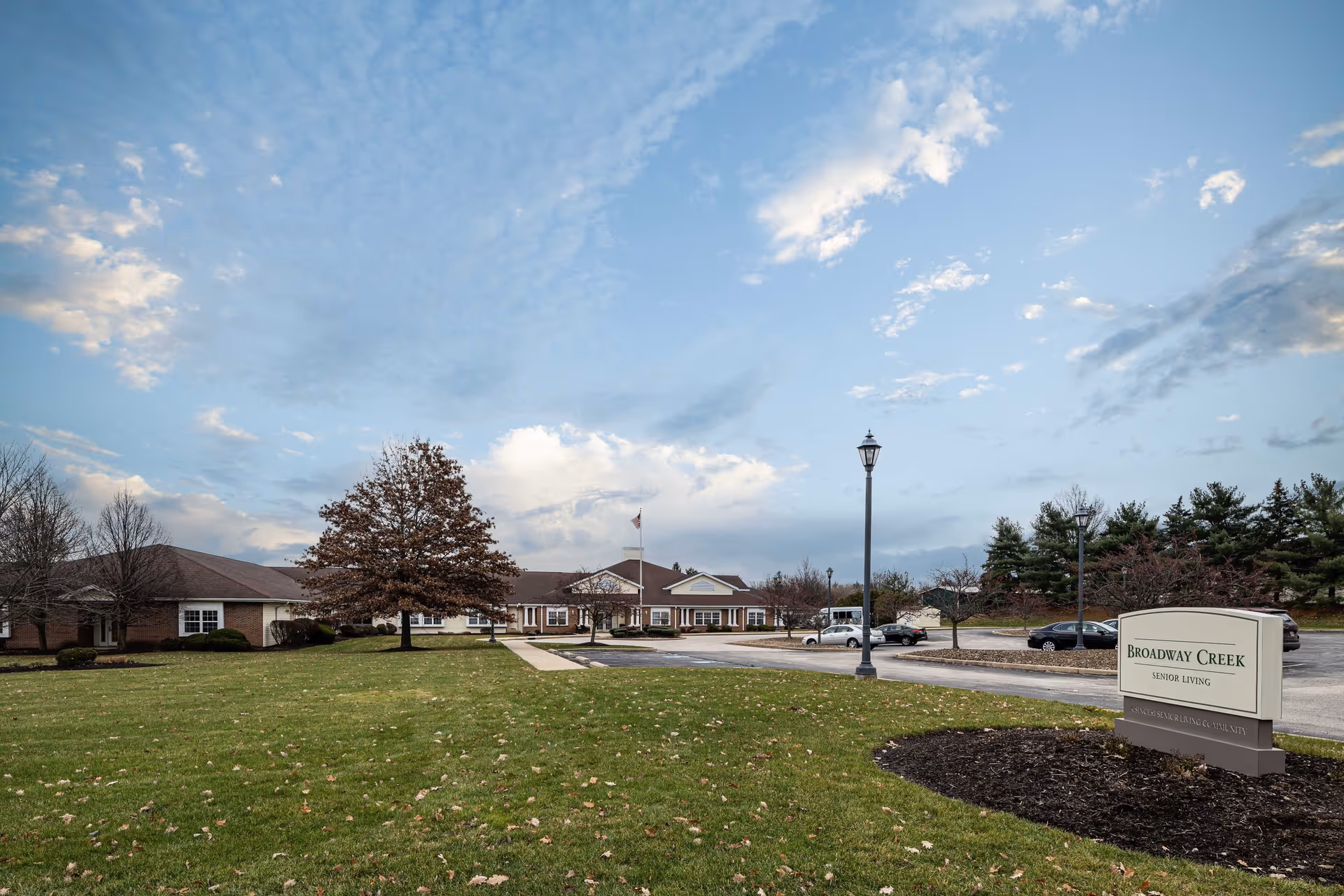 Exterior view of Broadway Creek Senior Living facility with a large lawn in the foreground, a few trees, a parking lot with several cars, and a sign displaying the facility's name. The sky is partly cloudy with a soft blue tone.