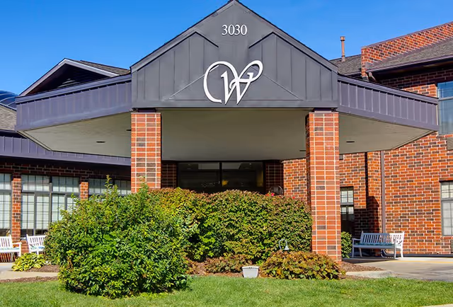 Front exterior view of Westgate Assisted Living building with brick walls, a covered entrance supported by brick columns, greenery and bushes in front, and a clear blue sky.