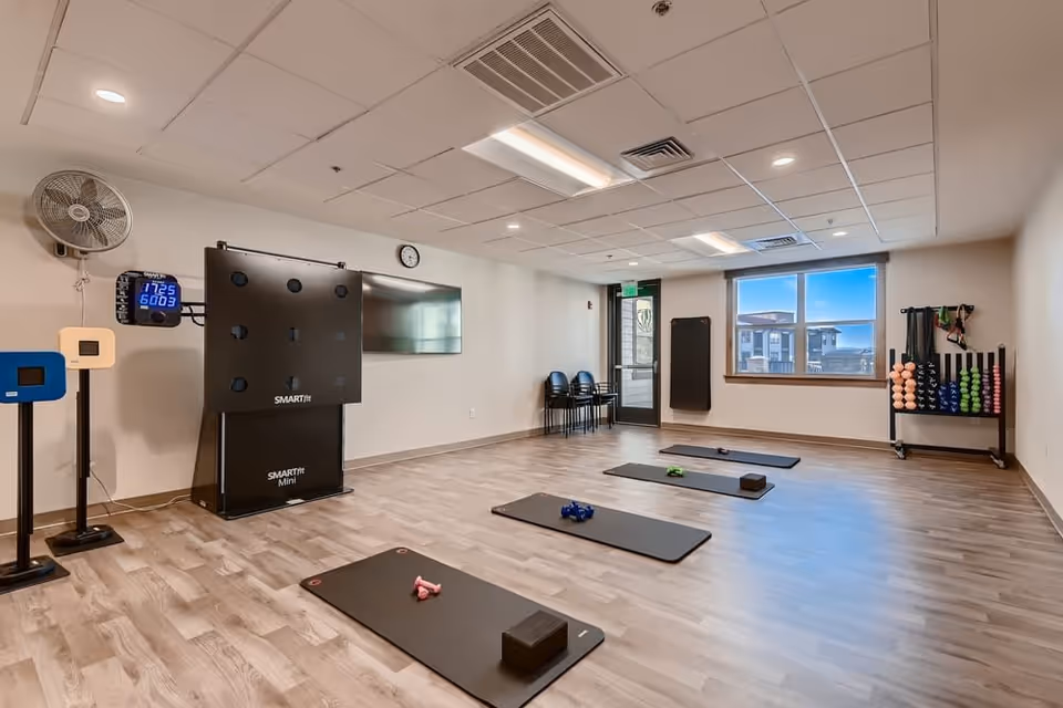 A bright multipurpose fitness room with yoga mats, dumbbells, weight racks, a wall-mounted screen, and a window.