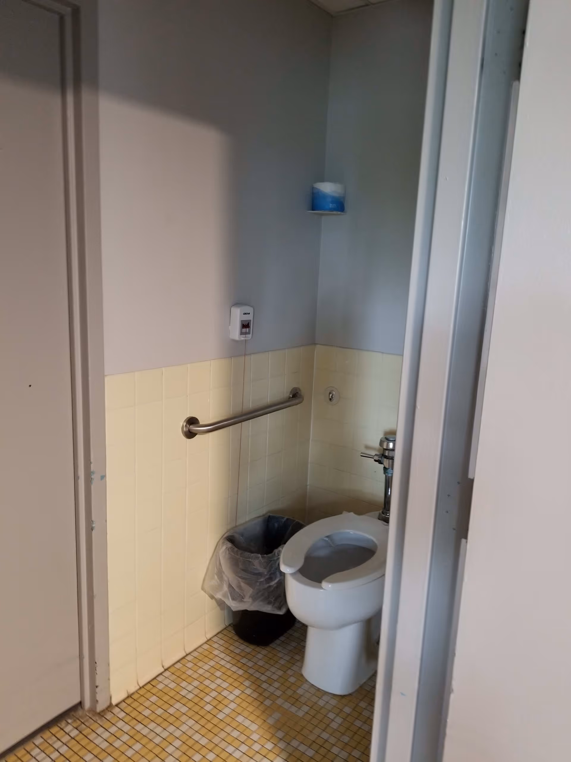 A small bathroom with a white toilet, a metal grab bar on the wall, a trash can with a plastic liner, a toilet paper roll on a small shelf, and beige tiled walls and floor.