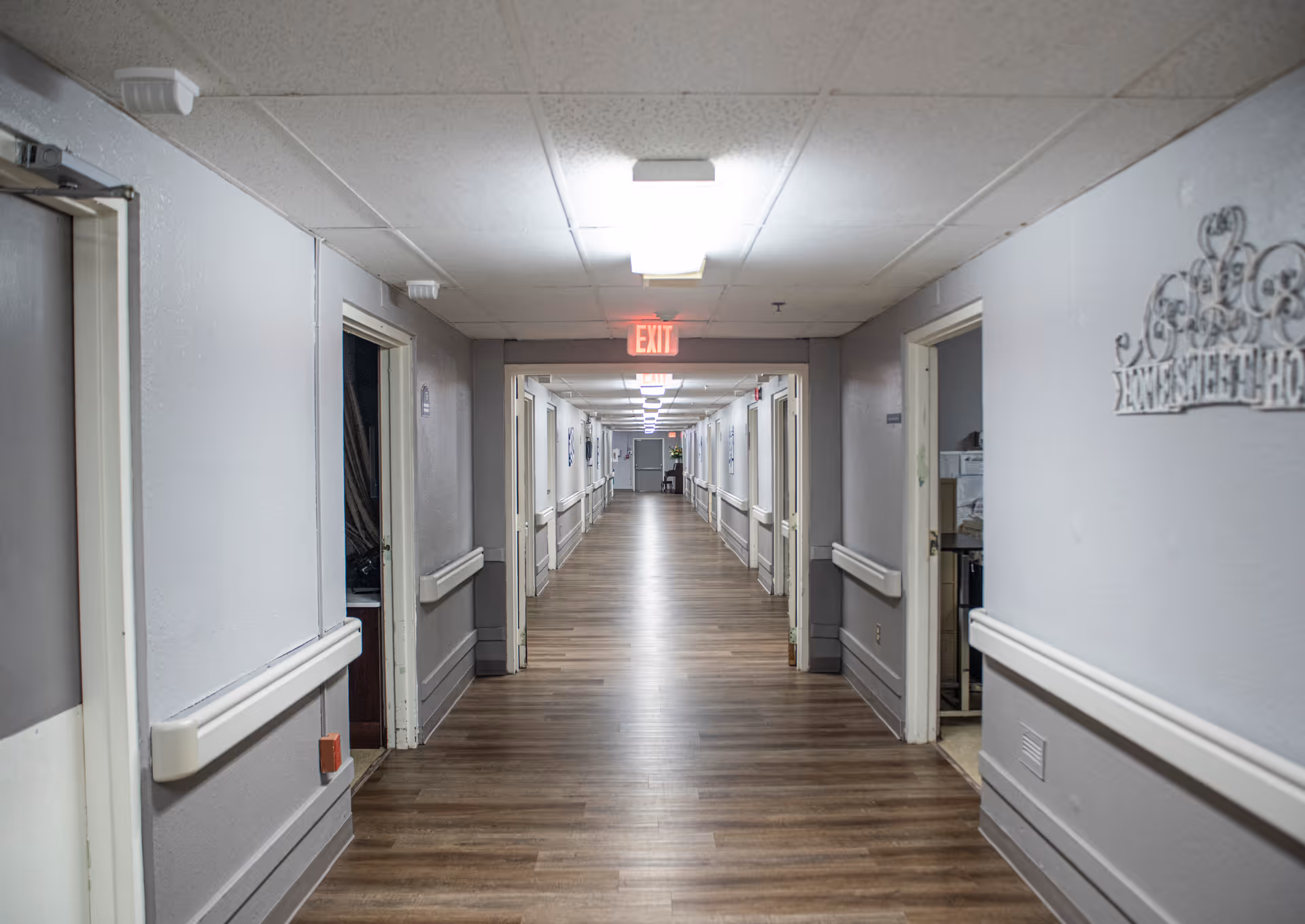 Long interior hallway of a senior living facility with wood flooring, handrails and doors on both sides and an EXIT sign at the far end.
