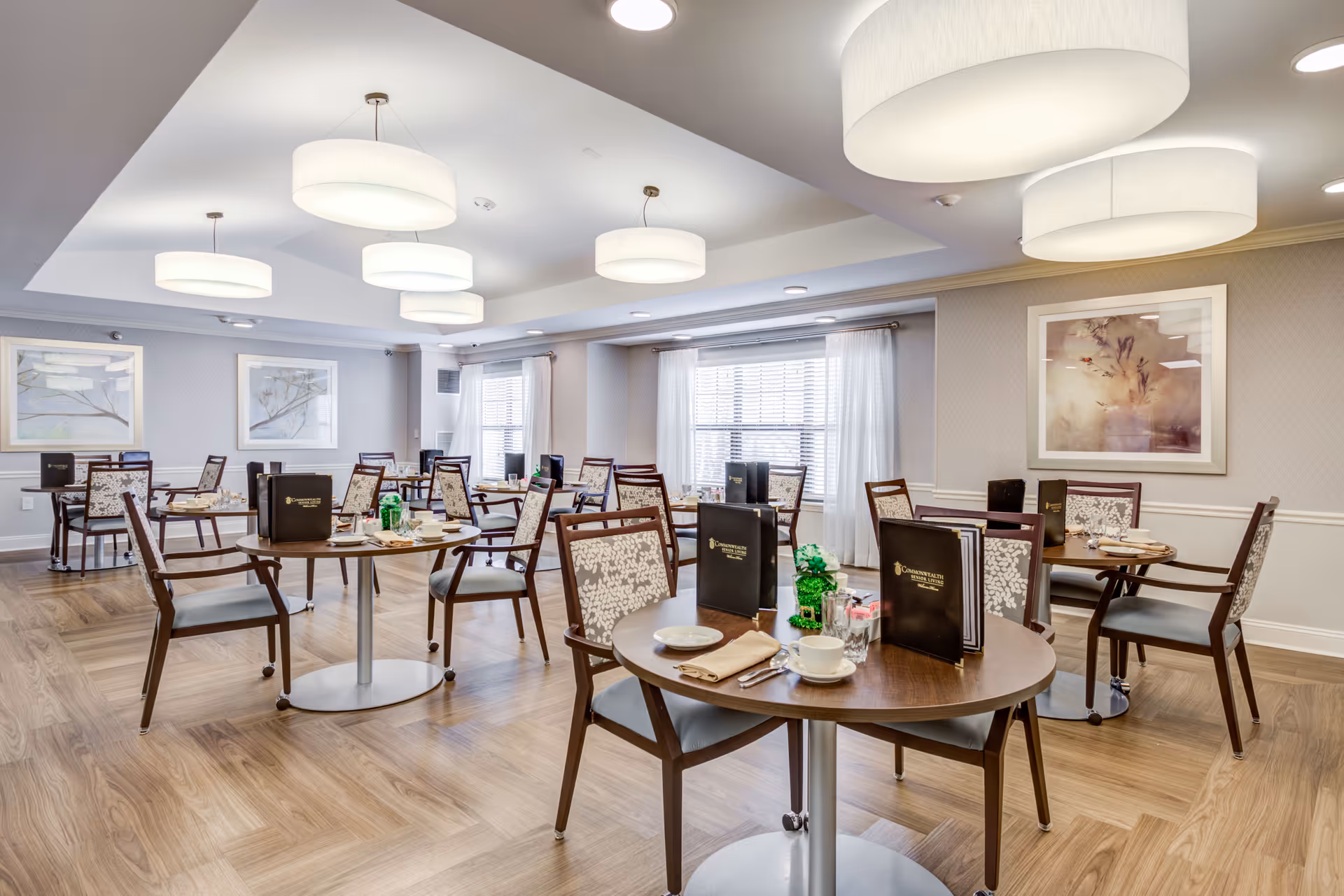 Well-lit senior living dining room with round tables set with place settings, chairs, menus, and large ceiling pendant lights.
