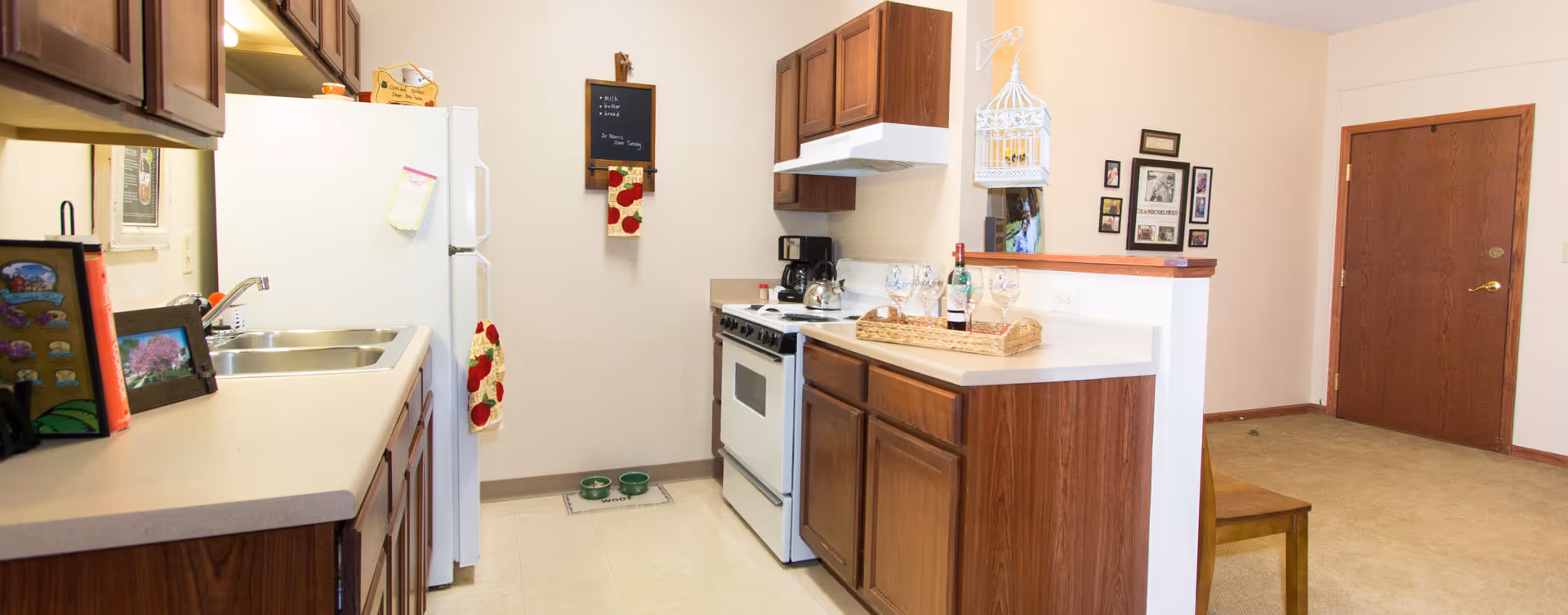 A kitchen area with wooden cabinets, a white refrigerator, a white stove, and a double sink. There are kitchen towels hanging on the refrigerator and wall, two pet bowls on the floor, a coffee maker on the counter, and a wine bottle with glasses on a tray. The kitchen opens to a carpeted area with a wooden door and a chair.