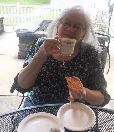 An elderly woman with white hair and glasses is sitting outdoors at a metal mesh table. She is holding a teacup in one hand and a piece of bread or pastry in the other. She is wearing a dark floral patterned blouse and a cross necklace. Behind her is a stone planter and a grassy area.