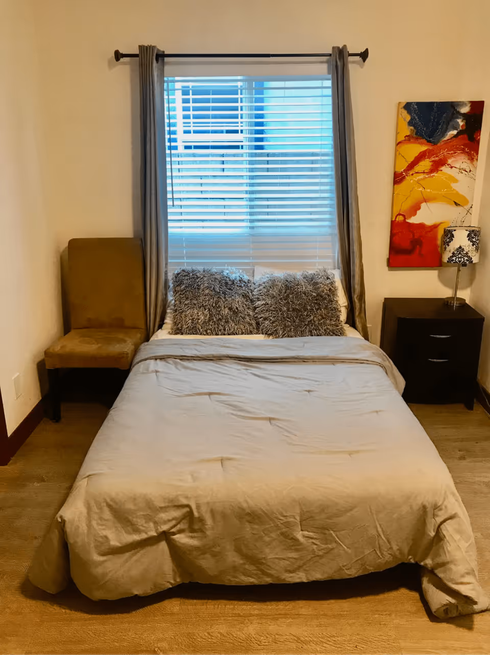 A neatly made bed with a beige comforter and two fluffy gray pillows positioned in front of a window with closed blinds and gray curtains. To the left of the bed is a brown upholstered chair, and to the right is a dark wooden nightstand with two drawers, topped with a decorative lamp and a colorful abstract painting on the wall above it. The room has wooden flooring and beige walls.