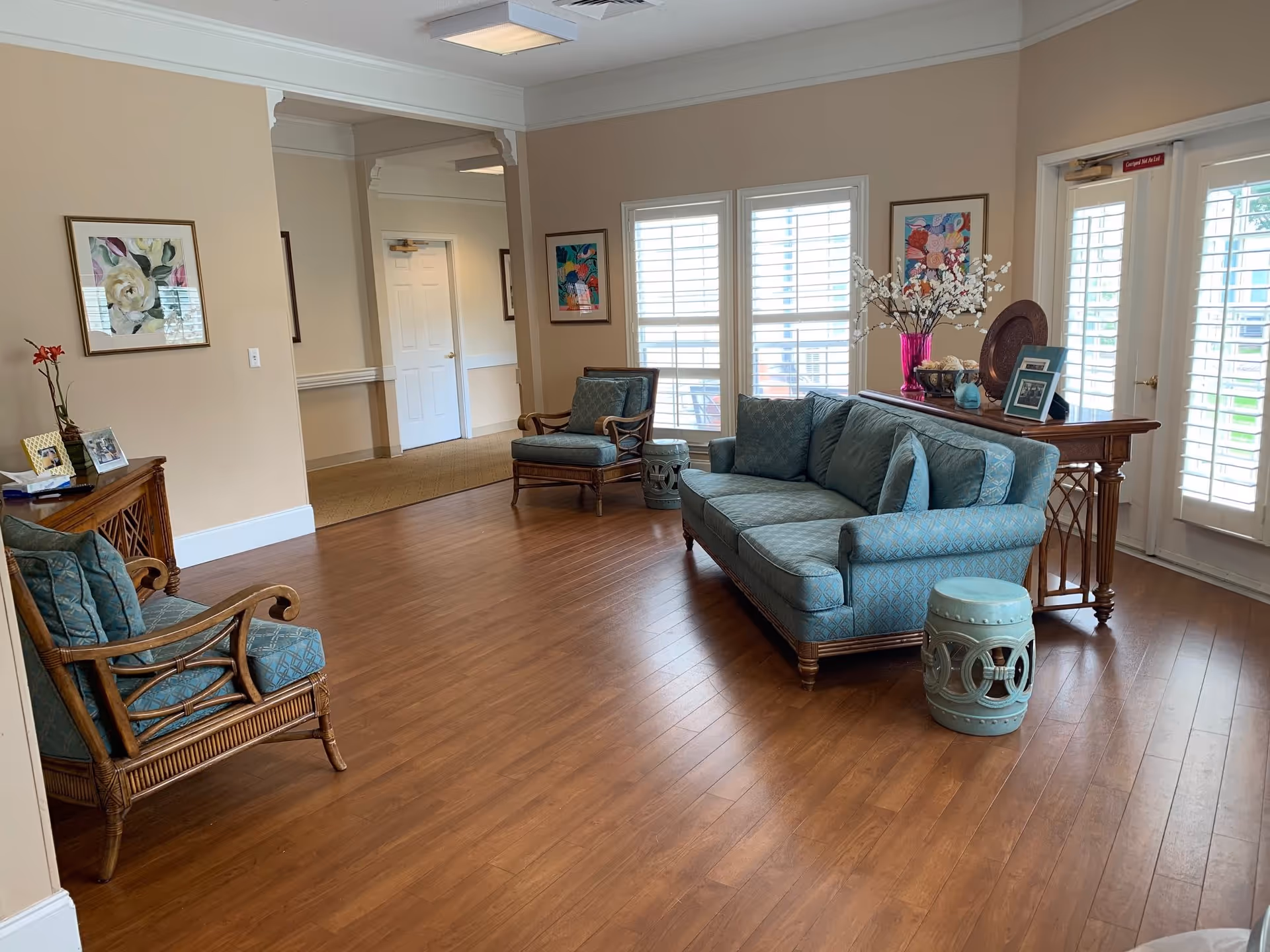 A cozy senior living facility common area with wooden flooring, a blue upholstered sofa, two matching armchairs, a small decorative stool, and a wooden console table adorned with framed photos and a vase of flowers. The room has beige walls, large windows with white shutters, and a glass door leading outside.