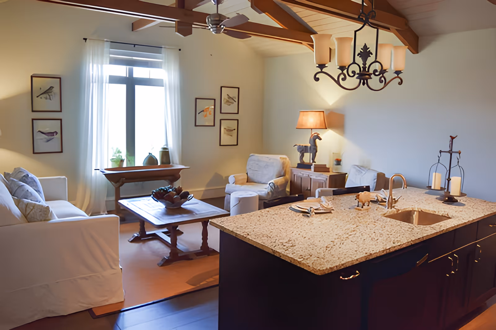 Open-plan living room and kitchen with a granite island in the foreground, seating area with sofa and armchairs, wooden ceiling beams and a window letting in natural light.