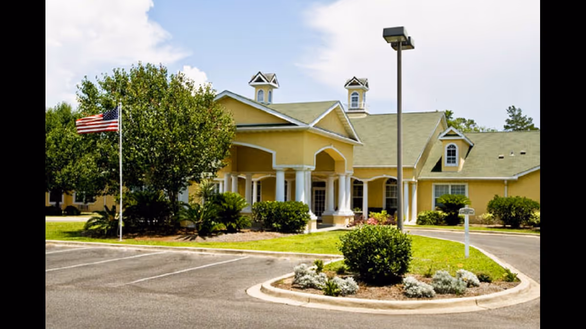 Exterior view of a yellow building with a green roof, featuring white columns at the entrance. There is a landscaped area with bushes and small plants in front, a flagpole with an American flag, and a parking lot in the foreground under a partly cloudy sky.