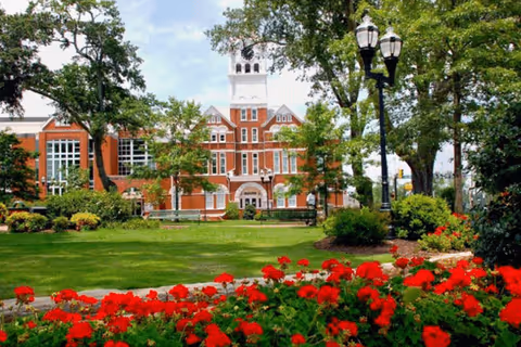 A large red brick building with a white clock tower surrounded by green trees and a well-maintained lawn with red flowers in the foreground under a partly cloudy sky.