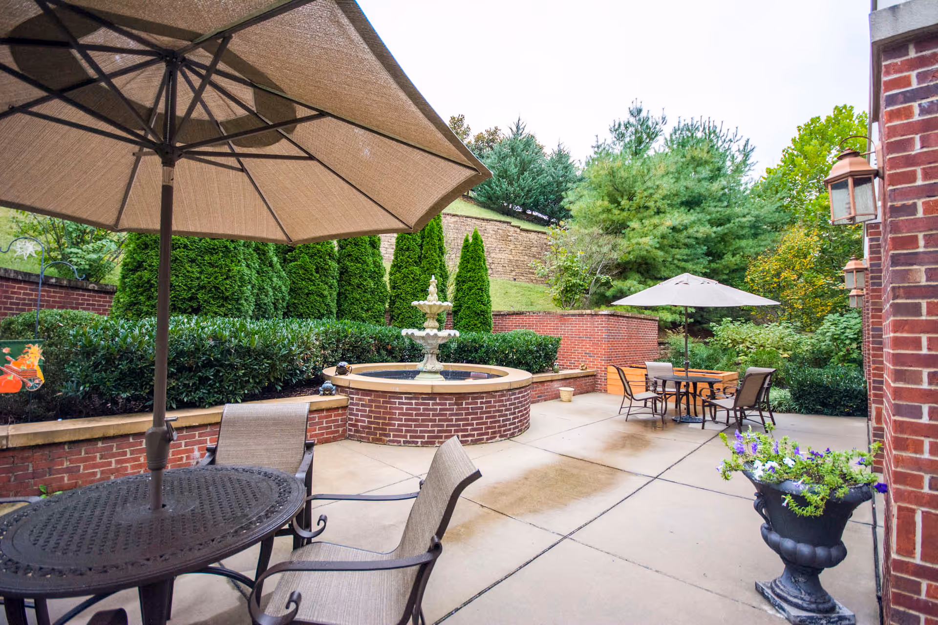 Outdoor brick courtyard with patio tables and umbrellas around a central tiered fountain and landscaped greenery.