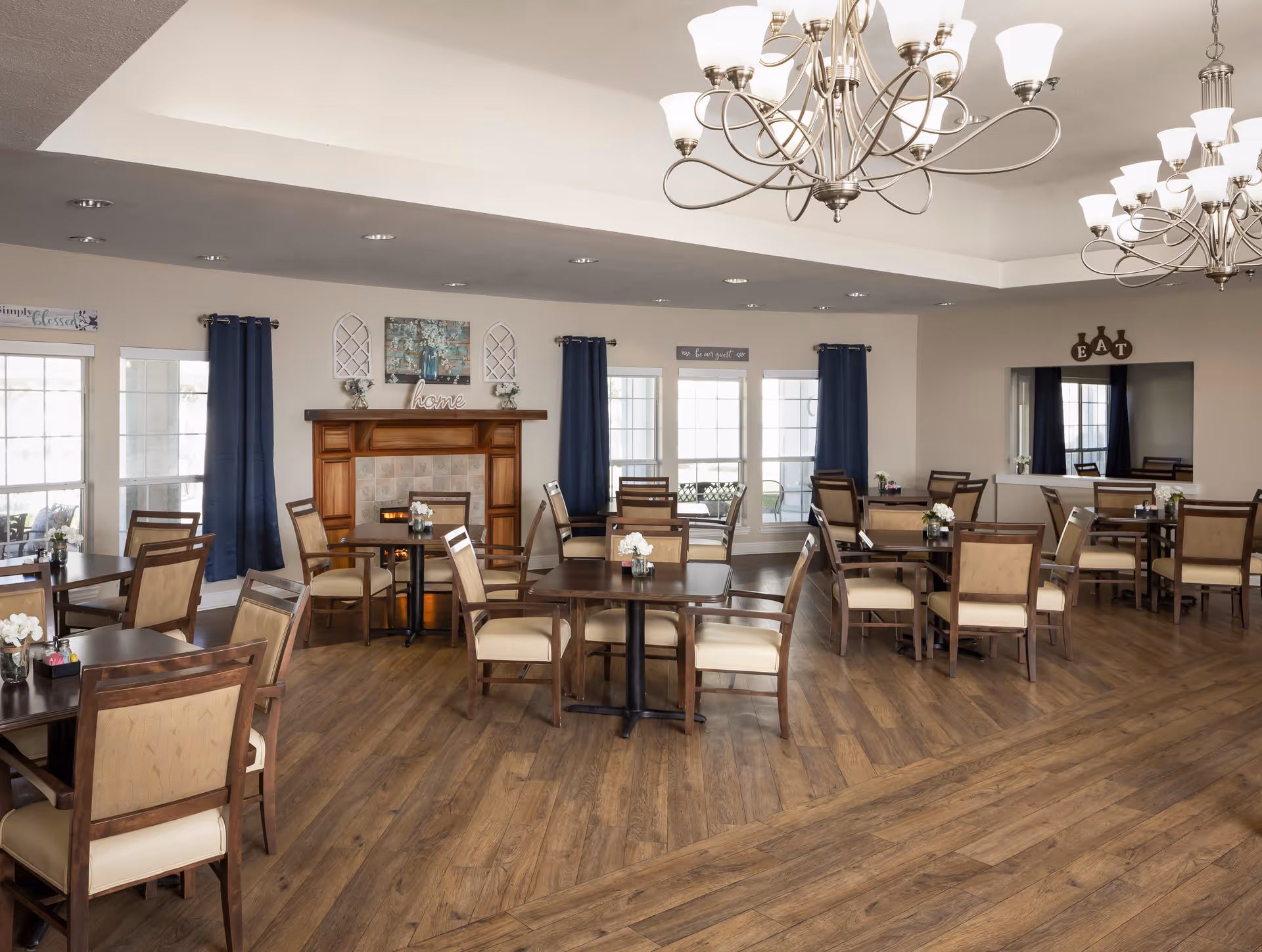 A spacious dining room with multiple wooden tables and cushioned chairs arranged neatly. The room features large windows with blue curtains, a decorative fireplace with the word 'home' on the mantel, and two elegant chandeliers hanging from the ceiling. The walls are light-colored, and there are small floral centerpieces on each table.