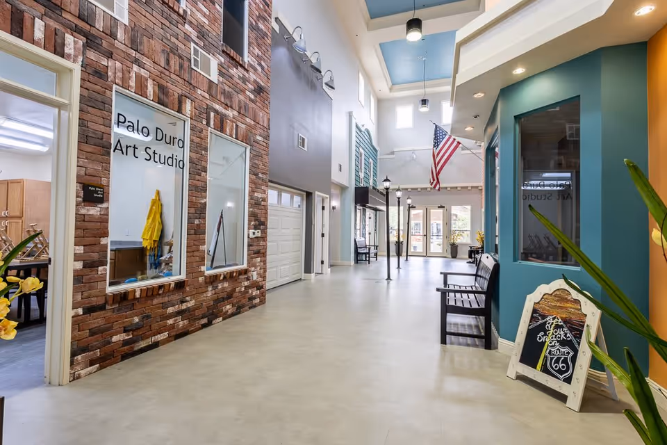 Interior hallway of Bristol Park at Amarillo Assisted & Living Memory Care featuring a high ceiling with skylights, an American flag hanging, benches along the walls, streetlamp-style lights, and a brick wall with windows labeled 'Palo Duro Art Studio'.