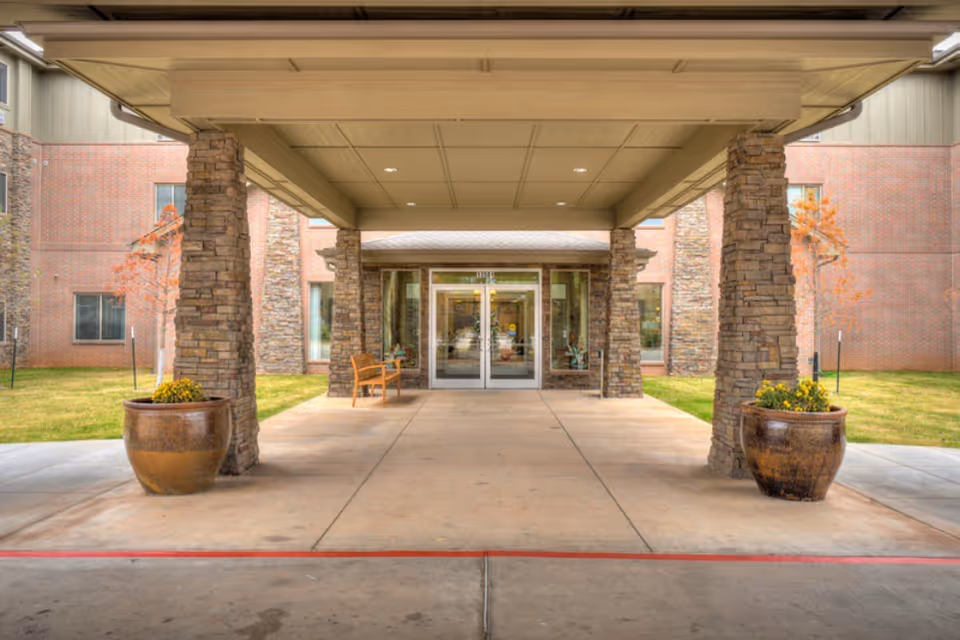 Covered entrance to a senior living facility with stone pillars, large planters with flowers, a bench, and glass double doors leading inside. The building exterior is brick with windows and some small trees on the lawn.