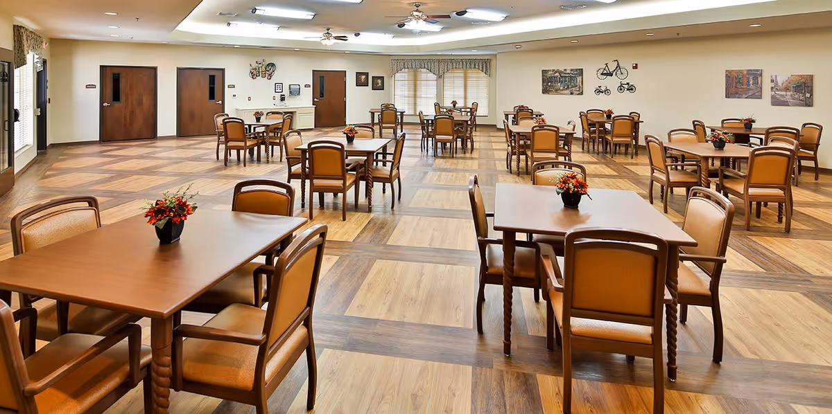 Spacious dining/activity room with multiple wooden tables and chairs and small floral centerpieces on a patterned wood floor.