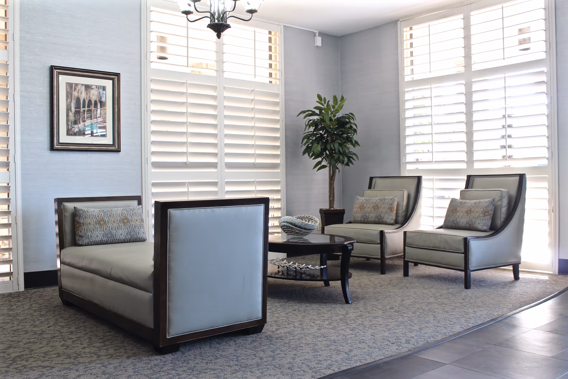 A bright and modern sitting area with two cushioned armchairs and a cushioned bench around a dark wooden coffee table. The room features large windows with white plantation shutters, a potted plant in the corner, a framed picture on the wall, and a chandelier hanging from the ceiling.