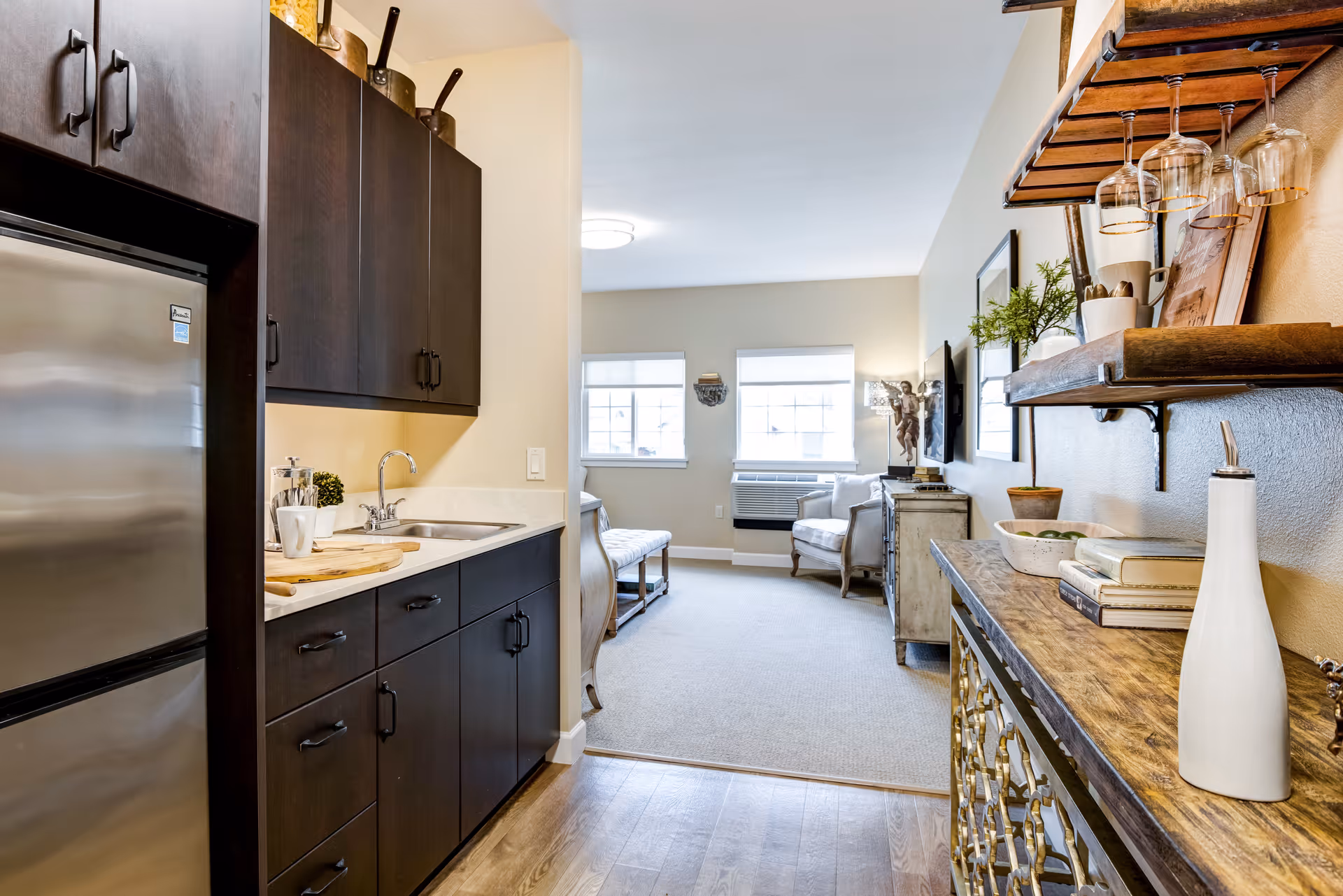 View of a small kitchen area with dark cabinets, a stainless steel refrigerator, and a sink on the left. The kitchen opens into a living room with beige walls, carpeted floor, two windows, a chair, and a TV mounted on the wall. On the right side, there is a wooden console table with decorative items and a hanging rack with wine glasses.