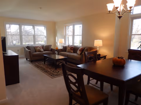 Open-plan living and dining area with sofas, a coffee table, a dining table, lamps, and large windows.