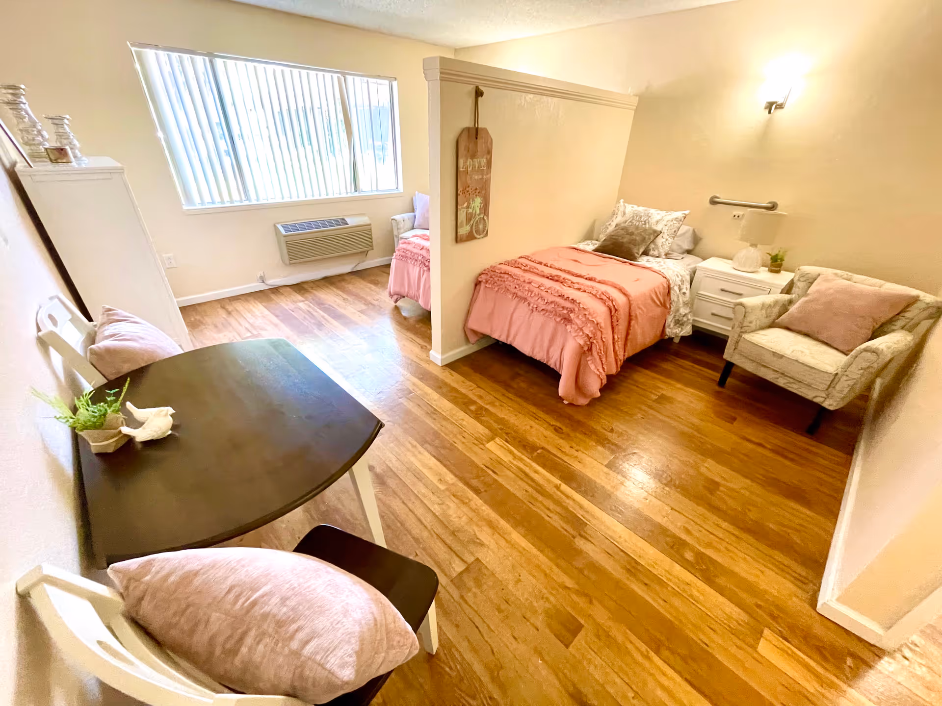 A cozy room with wooden flooring featuring two single beds separated by a half wall. Each bed has pink bedding and pillows. There is a white nightstand with a lamp and a small plant between the beds. A cushioned armchair with a pink pillow is placed next to one of the beds. Near the window, there is a small dark wooden table with two white chairs, each with a pink cushion. The room is well-lit with natural light coming through a large window with vertical blinds.