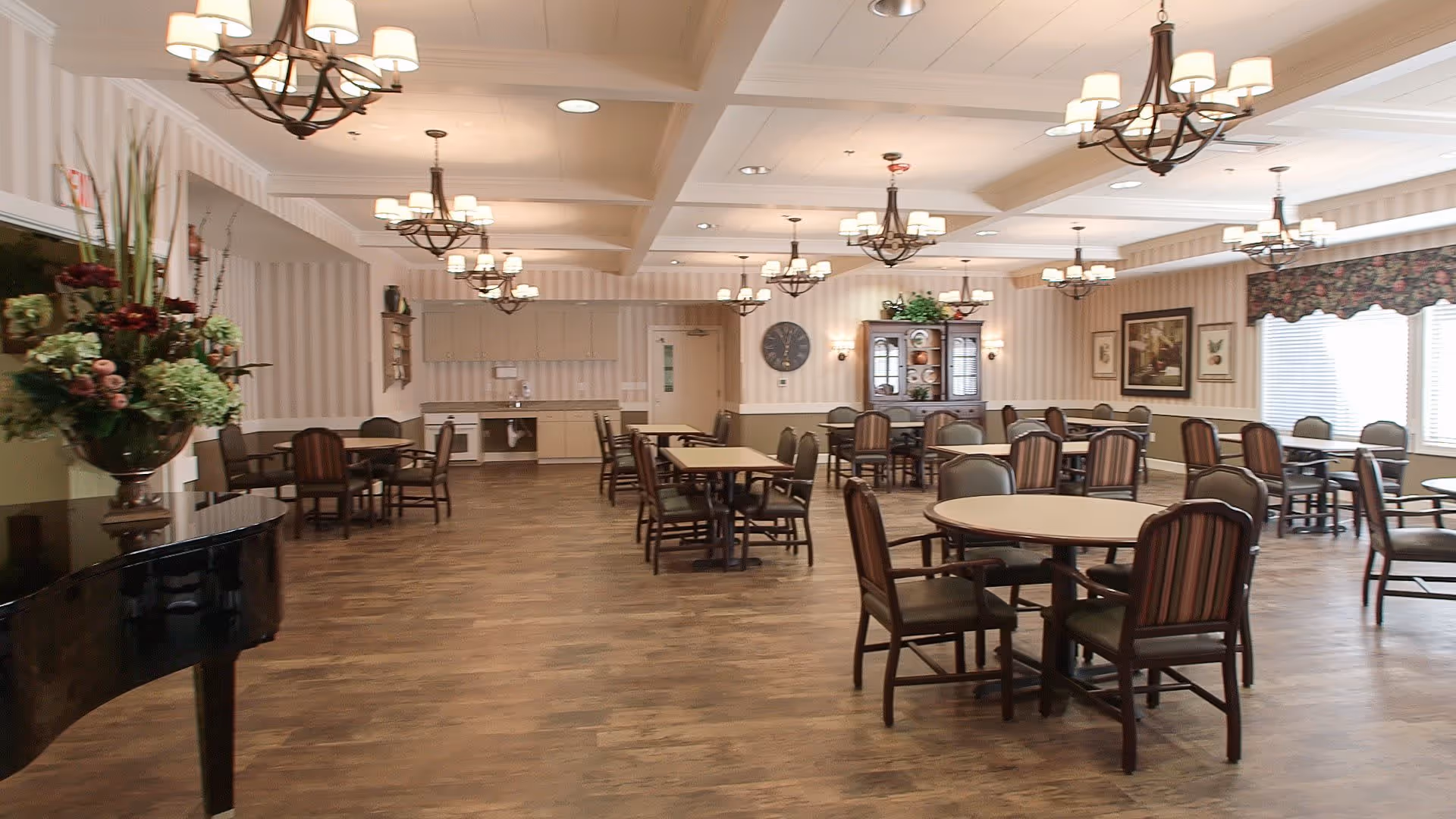 A spacious dining room in an assisted living facility with multiple round and rectangular tables surrounded by chairs. The room features wooden flooring, chandeliers hanging from the ceiling, a large floral arrangement on a black piano, and windows with floral valances allowing natural light to enter. The walls have striped wallpaper and framed artwork, and there is a cabinet with plants and decorative items.
