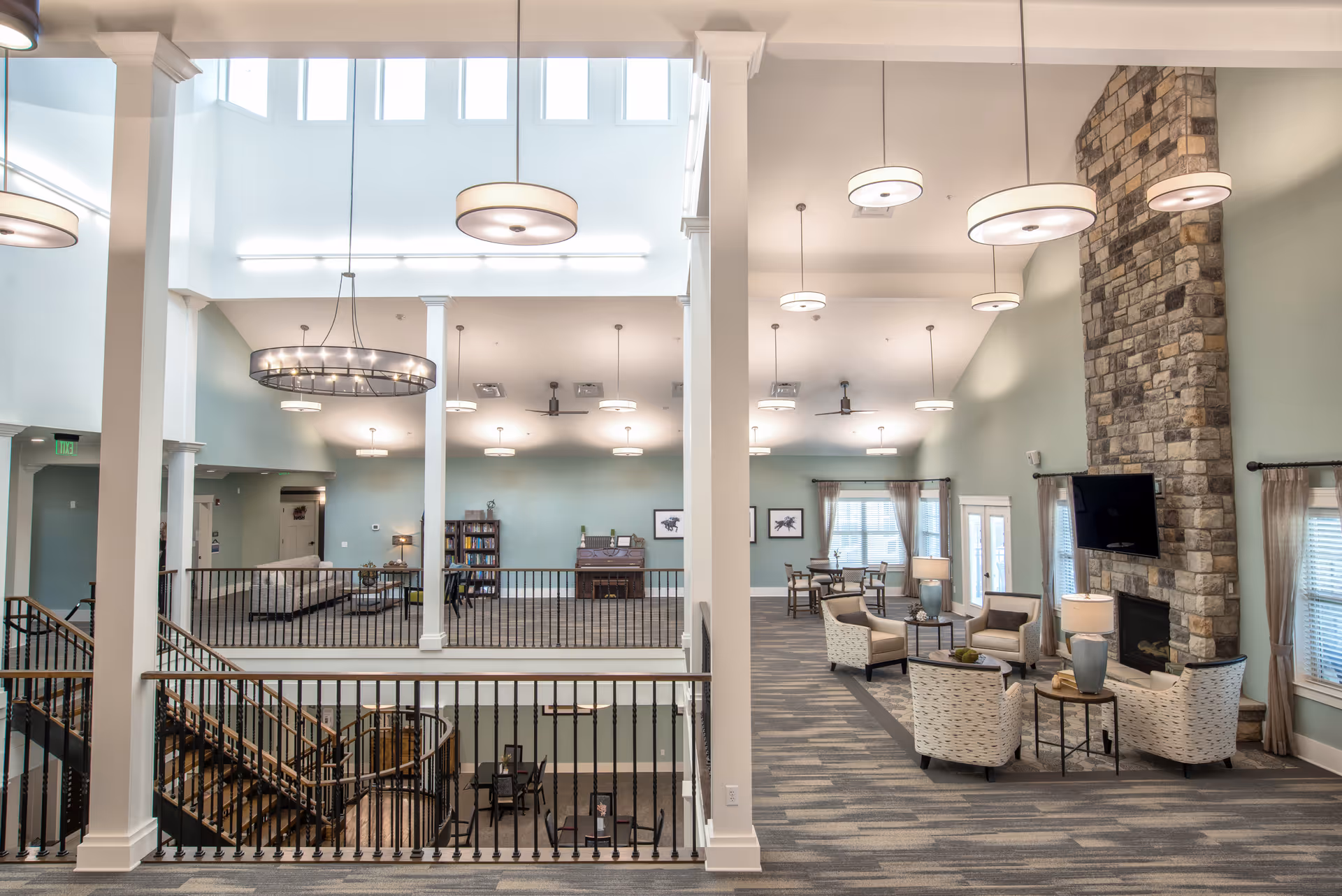 Spacious and well-lit senior living facility common area with high ceilings, modern hanging light fixtures, a stone fireplace with a mounted TV, comfortable seating arrangements including armchairs and sofas, a piano, bookshelves, and large windows with curtains allowing natural light.