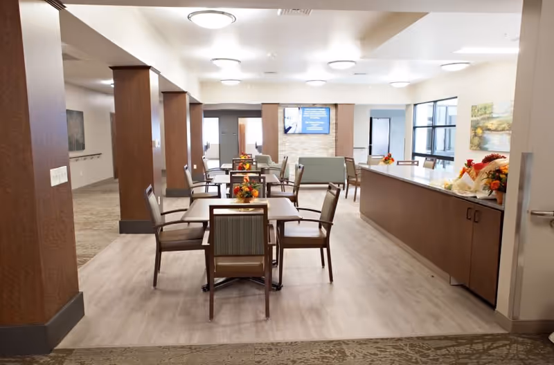A bright and spacious dining area in a senior living facility with several wooden tables and chairs arranged neatly. The room features light-colored flooring, wooden pillars, and a long counter on the right side decorated with floral arrangements. Large windows on the right wall allow natural light to enter, and a flat-screen TV is mounted on the far wall above a stone accent panel.