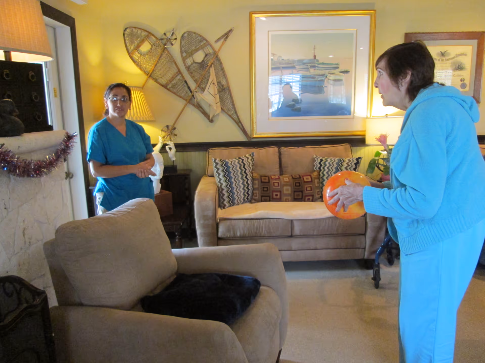 A senior woman in a light blue outfit holding an orange ball in a living room area, facing a caregiver in blue scrubs standing near a doorway. The room has beige furniture, decorative pillows, framed artwork on the walls, and a pair of snowshoes mounted on the wall.