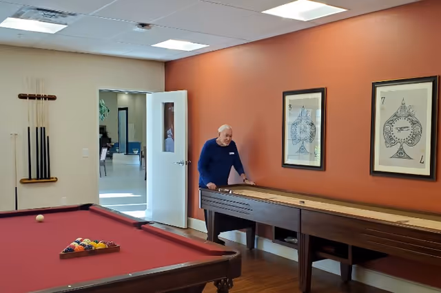 An elderly man playing shuffleboard in a recreational room with a pool table in the foreground. The room has a reddish-brown accent wall with two framed playing card artworks and a rack holding pool cues. A white door is open, revealing another room in the background.