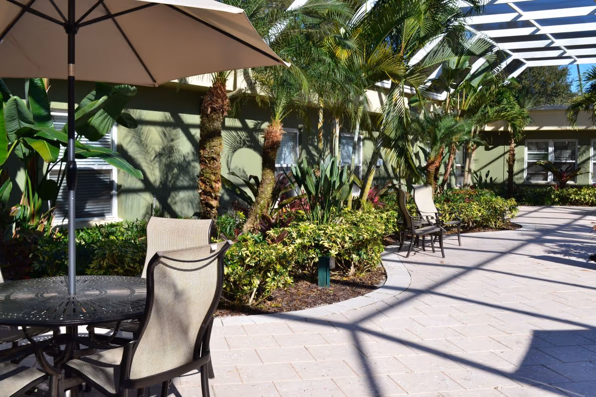 Outdoor patio area at Oak Manor Senior Living Community with a round table and chairs under a large beige umbrella, surrounded by lush green plants and palm trees, with a green building wall in the background and a pergola casting shadows on the paved ground.
