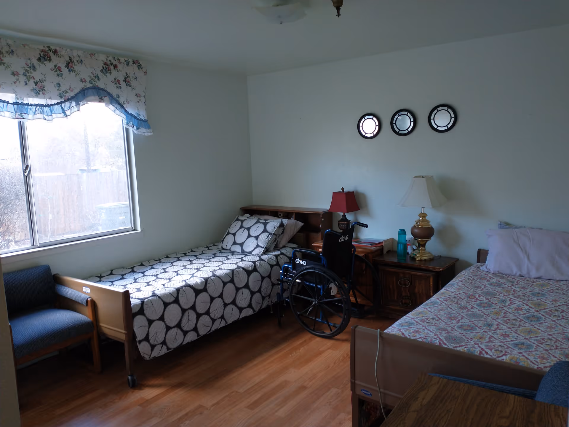 A senior living bedroom with two single beds, one with a black and white patterned bedspread and the other with a multicolored quilt. A wheelchair is positioned between the beds. There are two wooden nightstands with lamps and a water bottle on one. A window with floral curtains lets in natural light, and three decorative round mirrors hang on the wall above the beds. The floor is wooden, and there is a blue upholstered chair near the window.