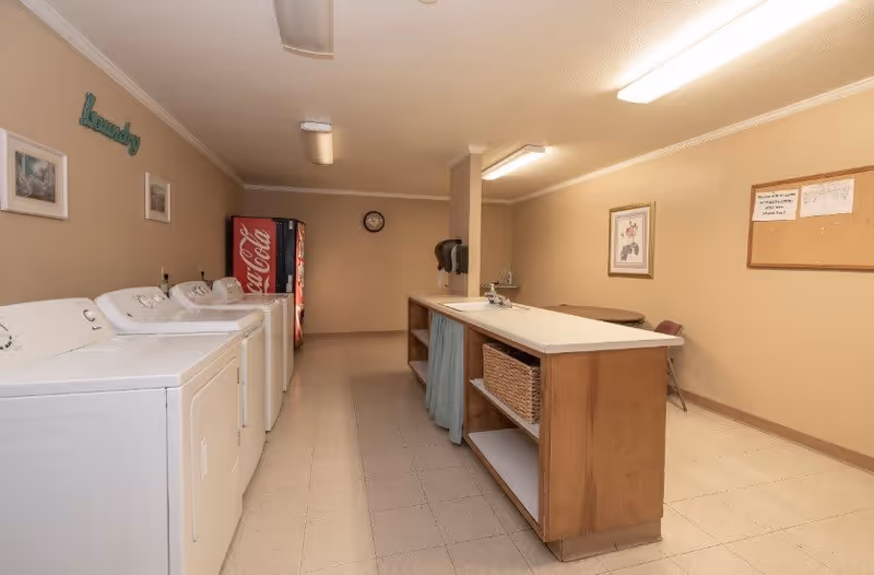 A laundry room in a senior living facility with three white washing machines and dryers lined up against the left wall. There is a wooden counter with a sink and shelves in the center of the room. A Coca-Cola vending machine is visible at the back, along with a clock on the wall. The walls are beige with framed pictures and a bulletin board. The word 'Laundry' is mounted on the left wall above the machines.