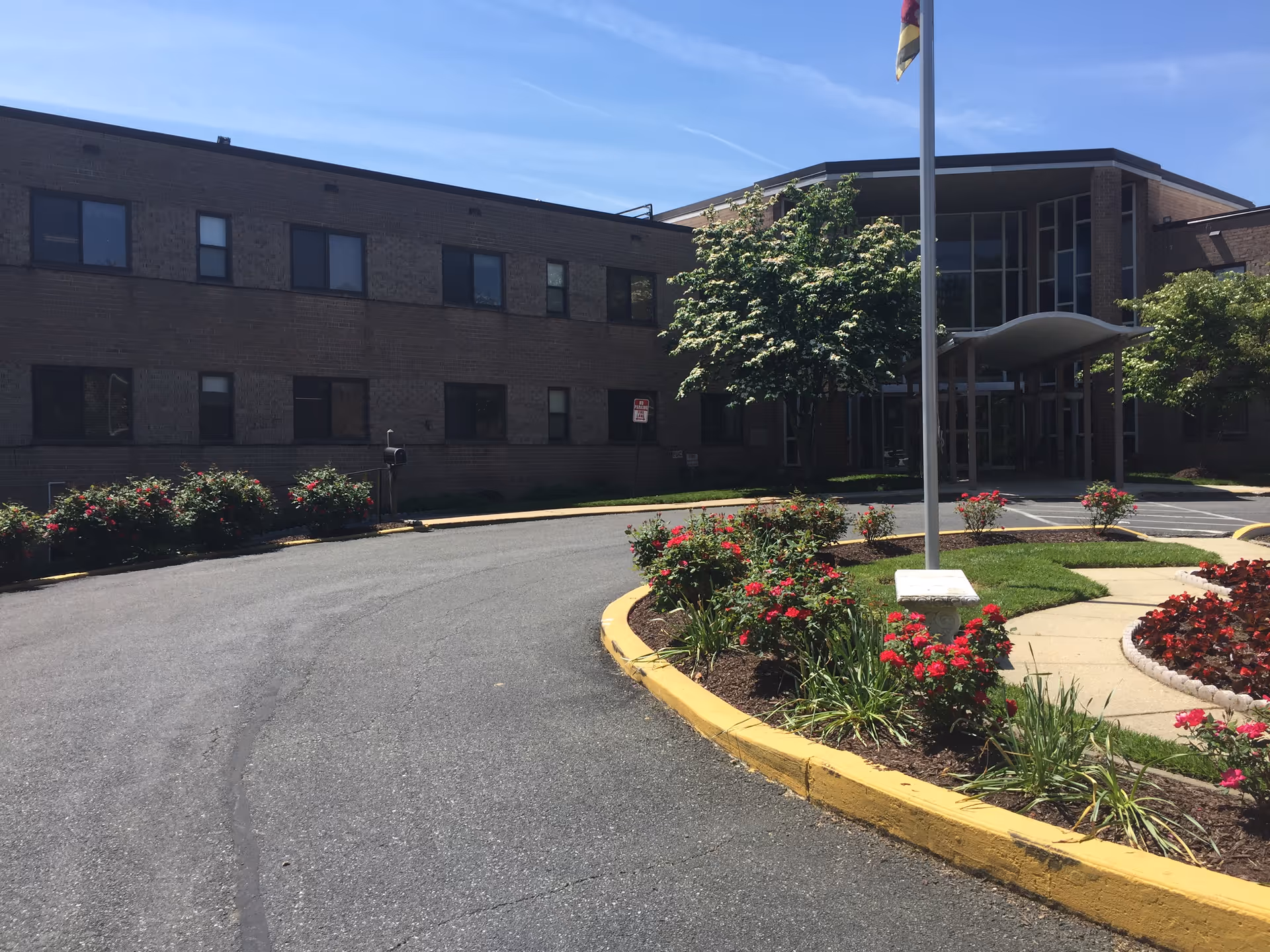 Front entrance of a brick nursing facility with a circular driveway, flagpole, and flower beds.