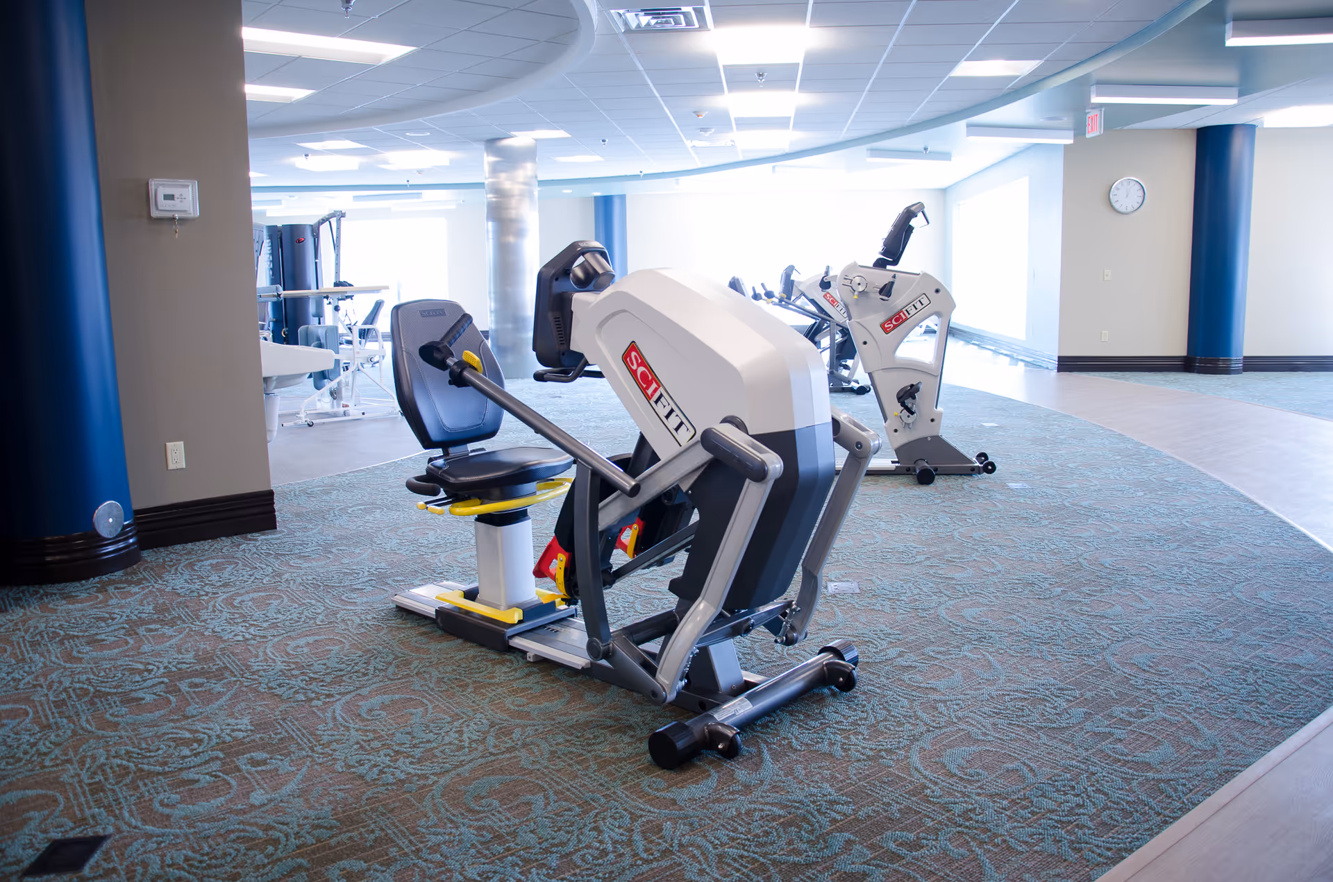 A bright and spacious rehabilitation gym area with exercise equipment including a SciFit machine on patterned carpet flooring. The room has a curved ceiling design with recessed lighting, large windows letting in natural light, and a clock on the wall.