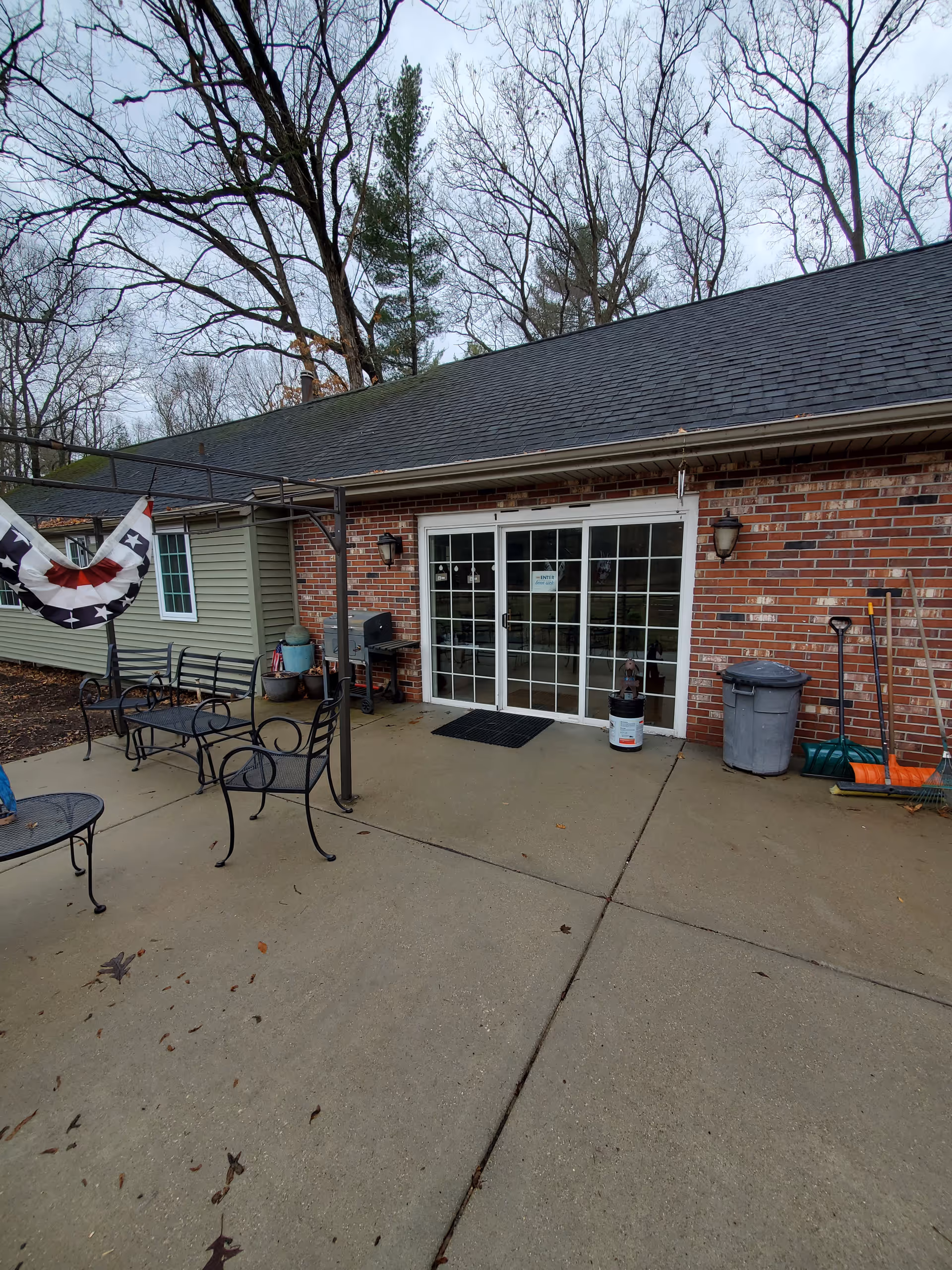 Outdoor patio area with metal chairs and tables on a concrete surface in front of a brick building with sliding glass doors. There are some gardening tools, a trash can, and a grill near the building. Leafless trees are visible in the background under a cloudy sky.