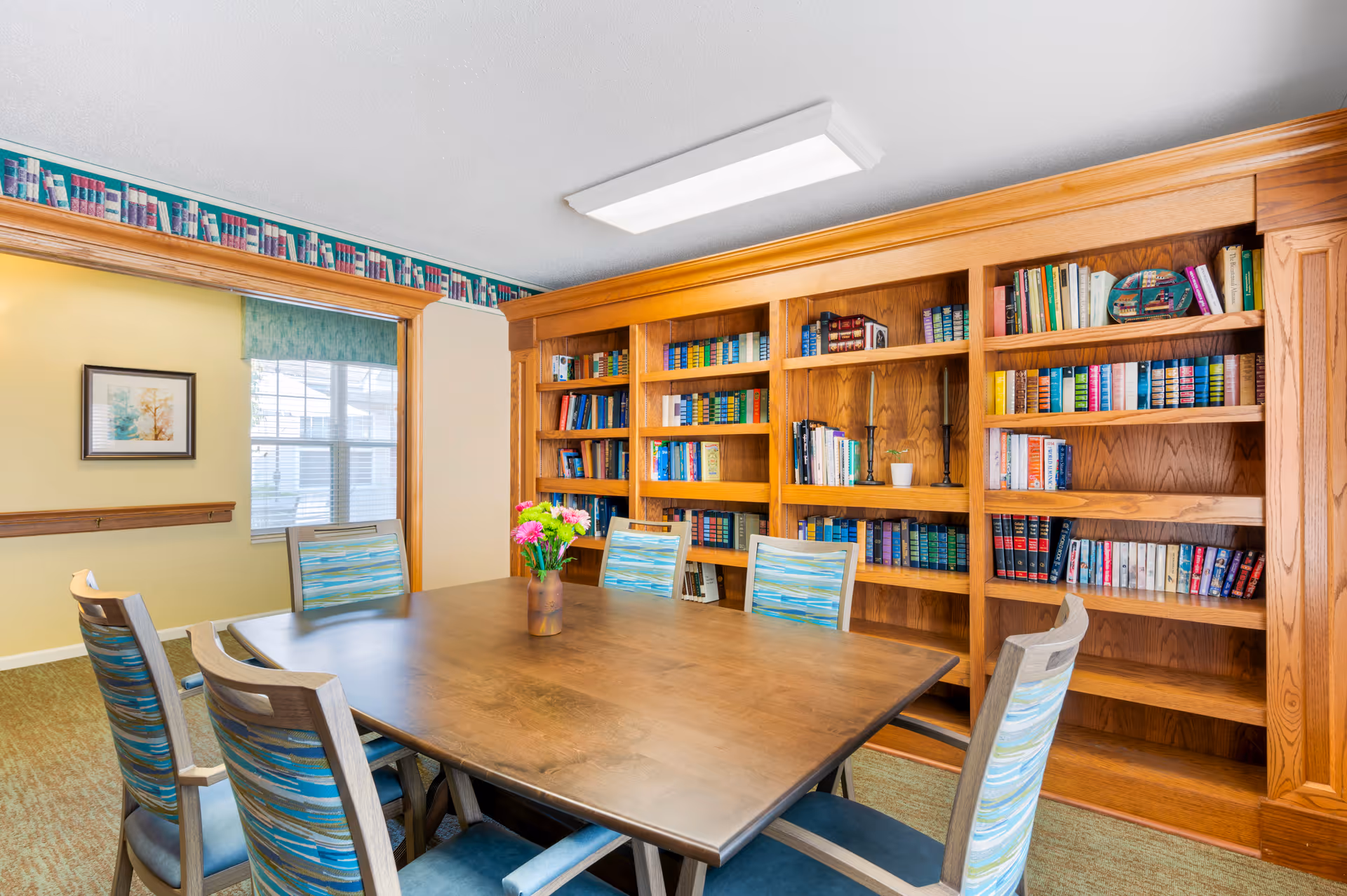 A well-lit common room with a wooden table surrounded by striped chairs and a large built-in bookshelf filled with books.