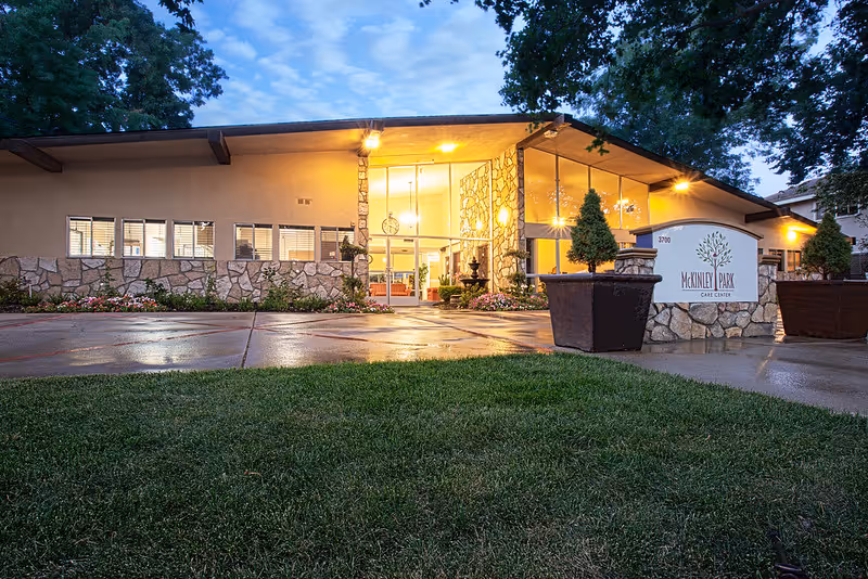Exterior view of McKinley Park Care Center building at dusk, showing a well-lit entrance with large windows, stone accents, and a sign with the facility's name. There is a green lawn and large potted plants near the entrance.