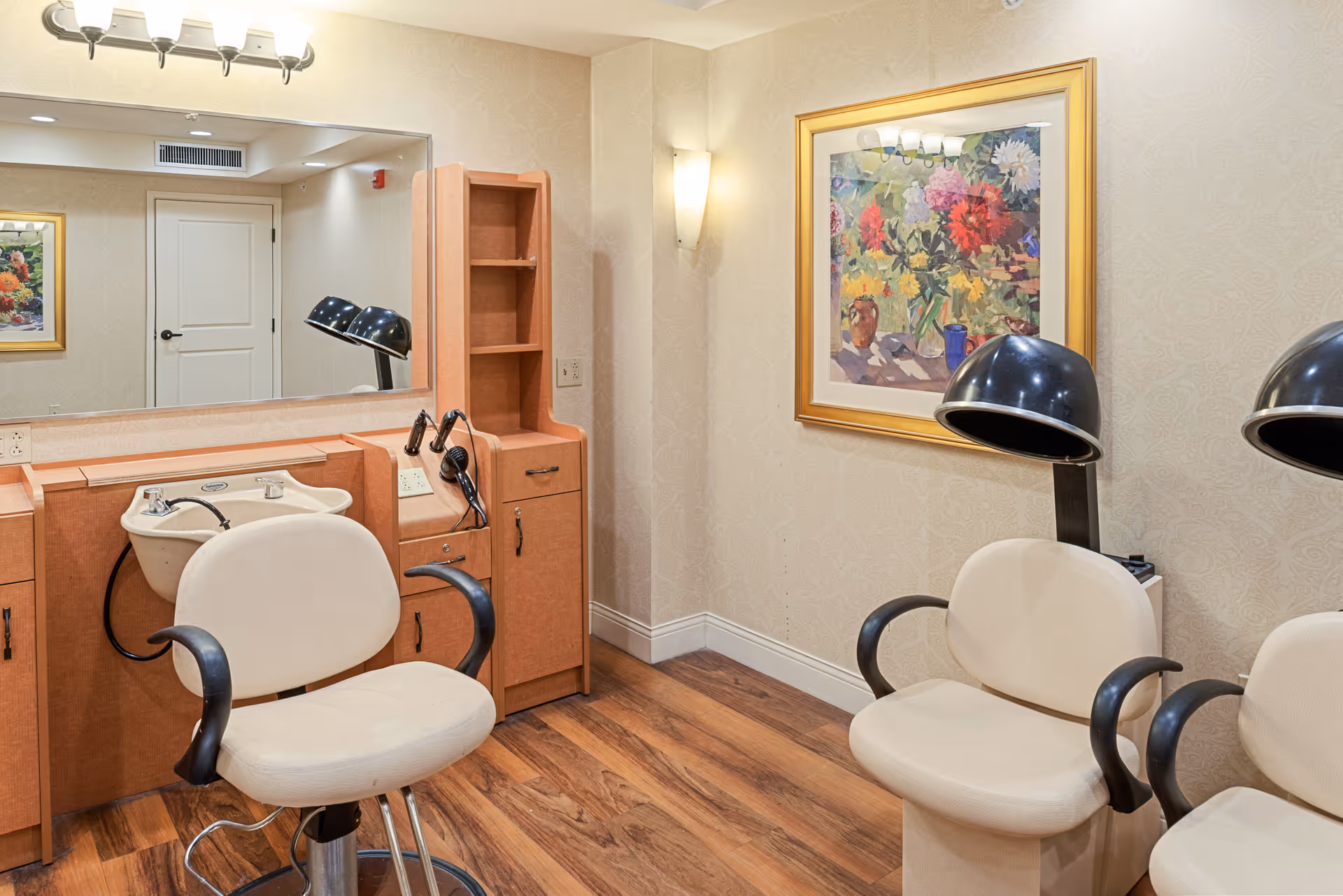Small salon room with styling chairs, hooded hair dryers, a shampoo sink, large mirror, and framed artwork.