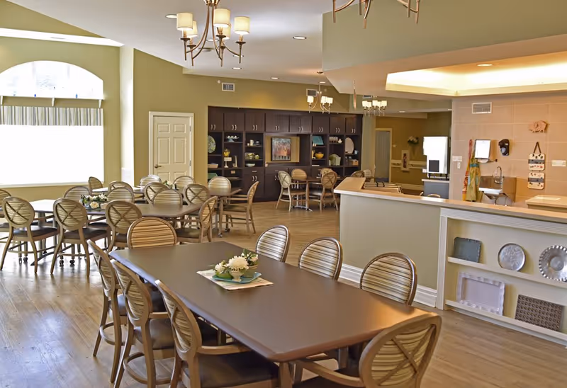 A spacious dining room in an assisted living facility with multiple tables and chairs arranged neatly. The room features wooden flooring, a large window with a valance, decorative light fixtures on the ceiling, and a built-in cabinet with shelves displaying various items. There is also a counter area with decorative plates on the side and a small sink on the wall.