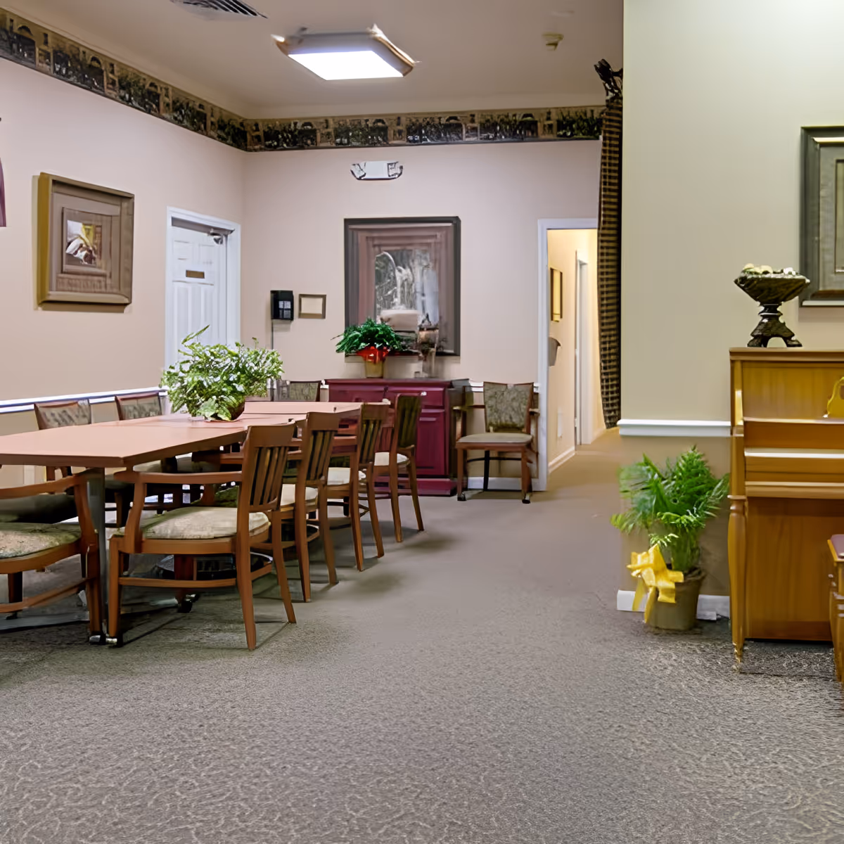 A dining room with a long wooden table surrounded by chairs, a potted plant on the table, a piano on the right side with a plant next to it, framed artwork on the walls, and a hallway leading to other rooms.