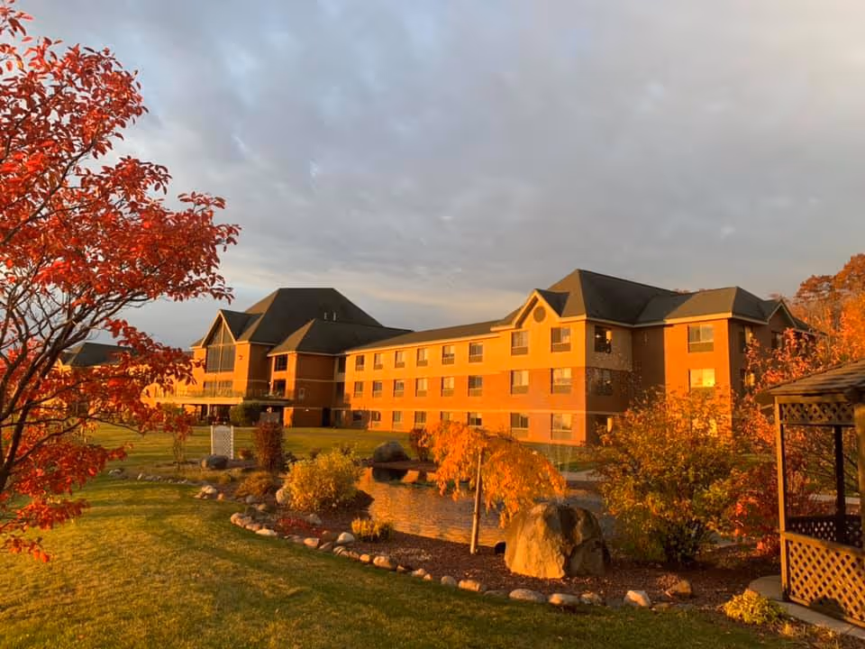 Three-story senior living building set behind a landscaped pond and autumn trees under a cloudy sunset sky.