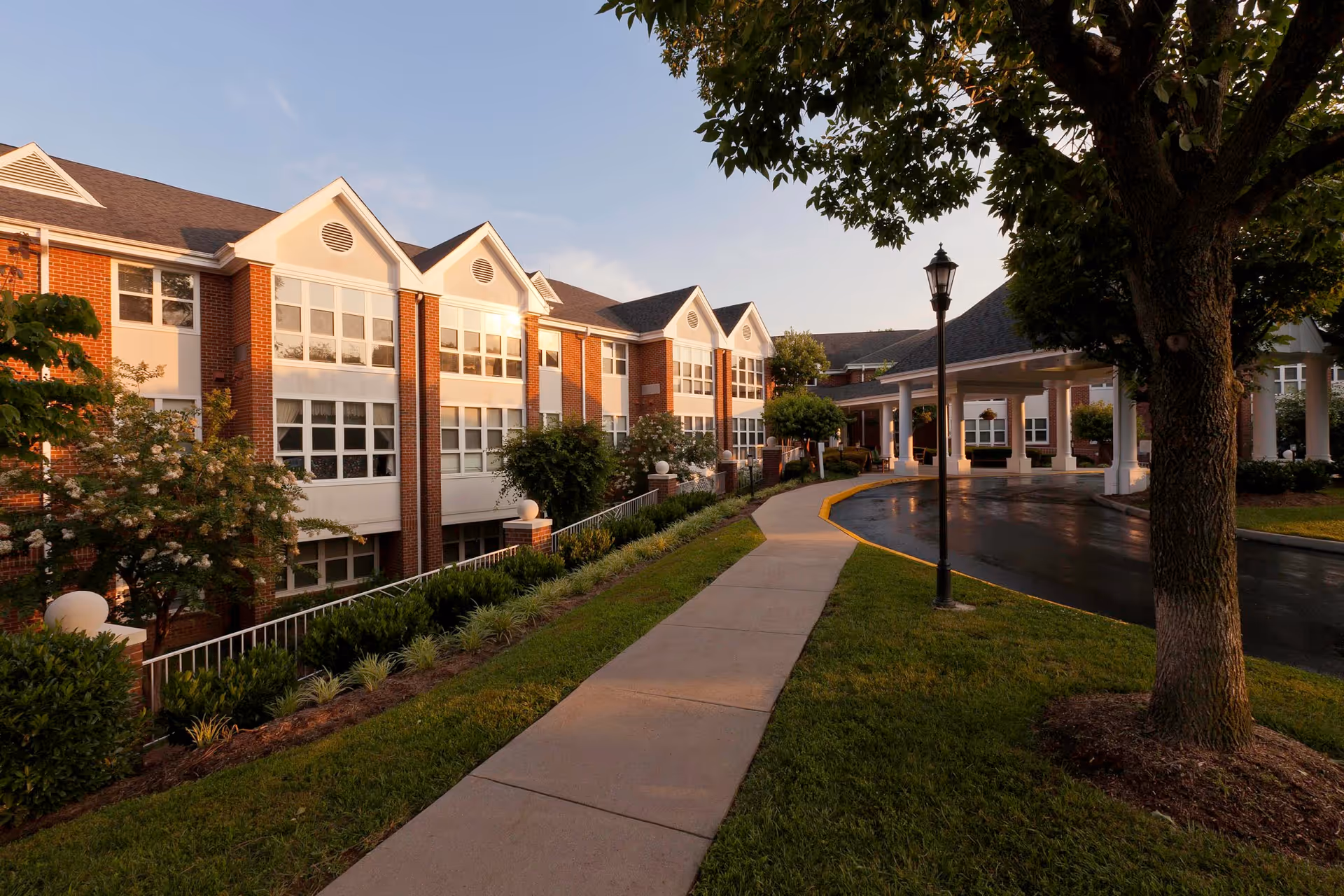 Exterior view of a senior living facility named Our Lady of Peace during sunset, showing a brick building with large windows, a covered entrance, a sidewalk, green grass, trees, and a street lamp.