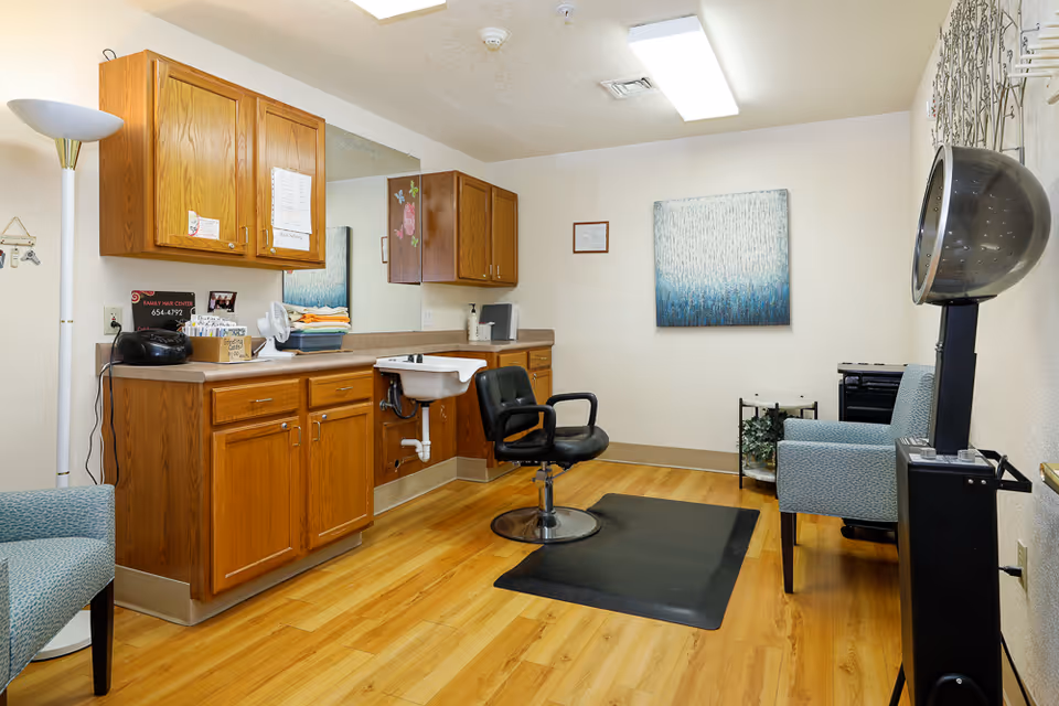 Interior of a hair salon room in an assisted living facility featuring wooden cabinets, a sink, a black salon chair on a black mat, two upholstered chairs, a hair dryer hood, a floor lamp, and a blue and white abstract painting on the wall.