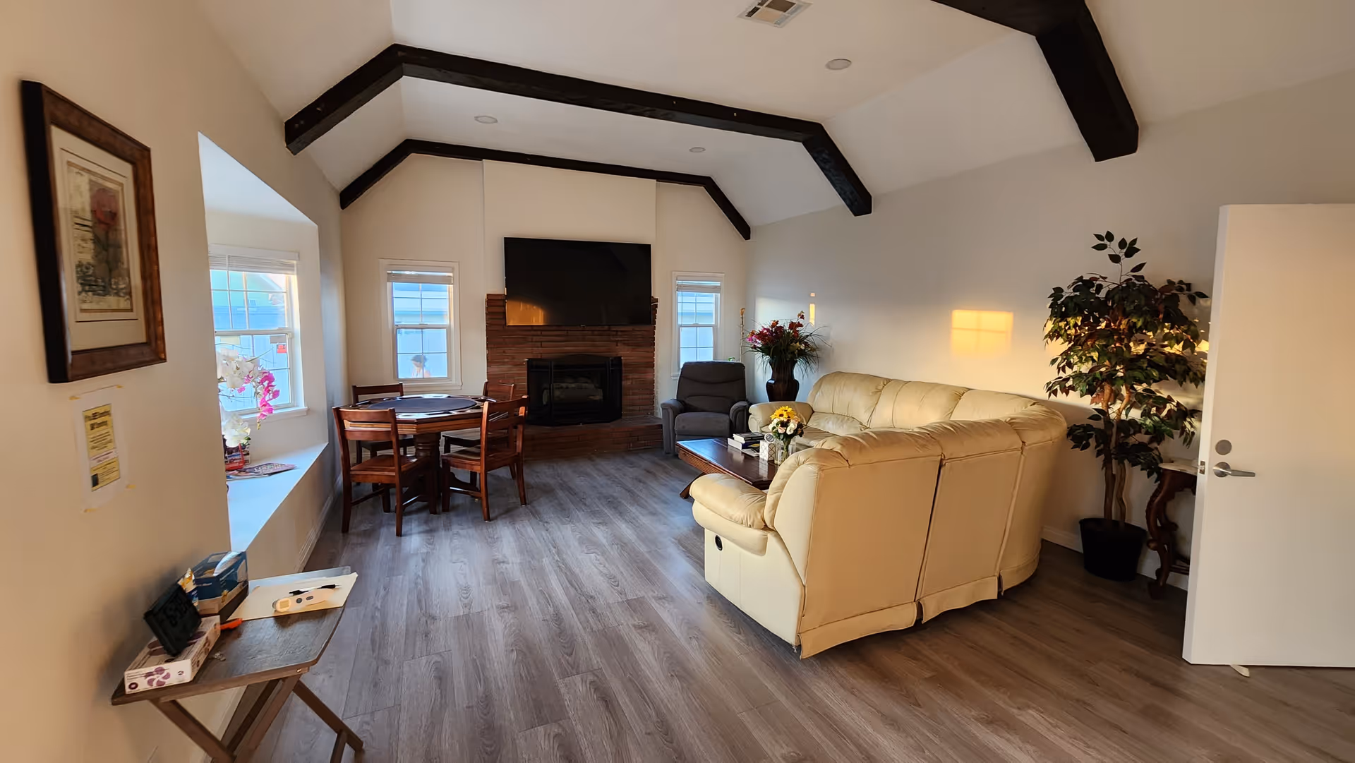 A cozy senior living room with a beige leather sectional sofa, a dark armchair, a wooden coffee table with flowers, and a round wooden dining table with four chairs. The room features a fireplace with a mounted flat-screen TV above it, two windows on either side of the fireplace, wooden ceiling beams, and light wood flooring. There is a potted plant near an open door and framed artwork on the wall.
