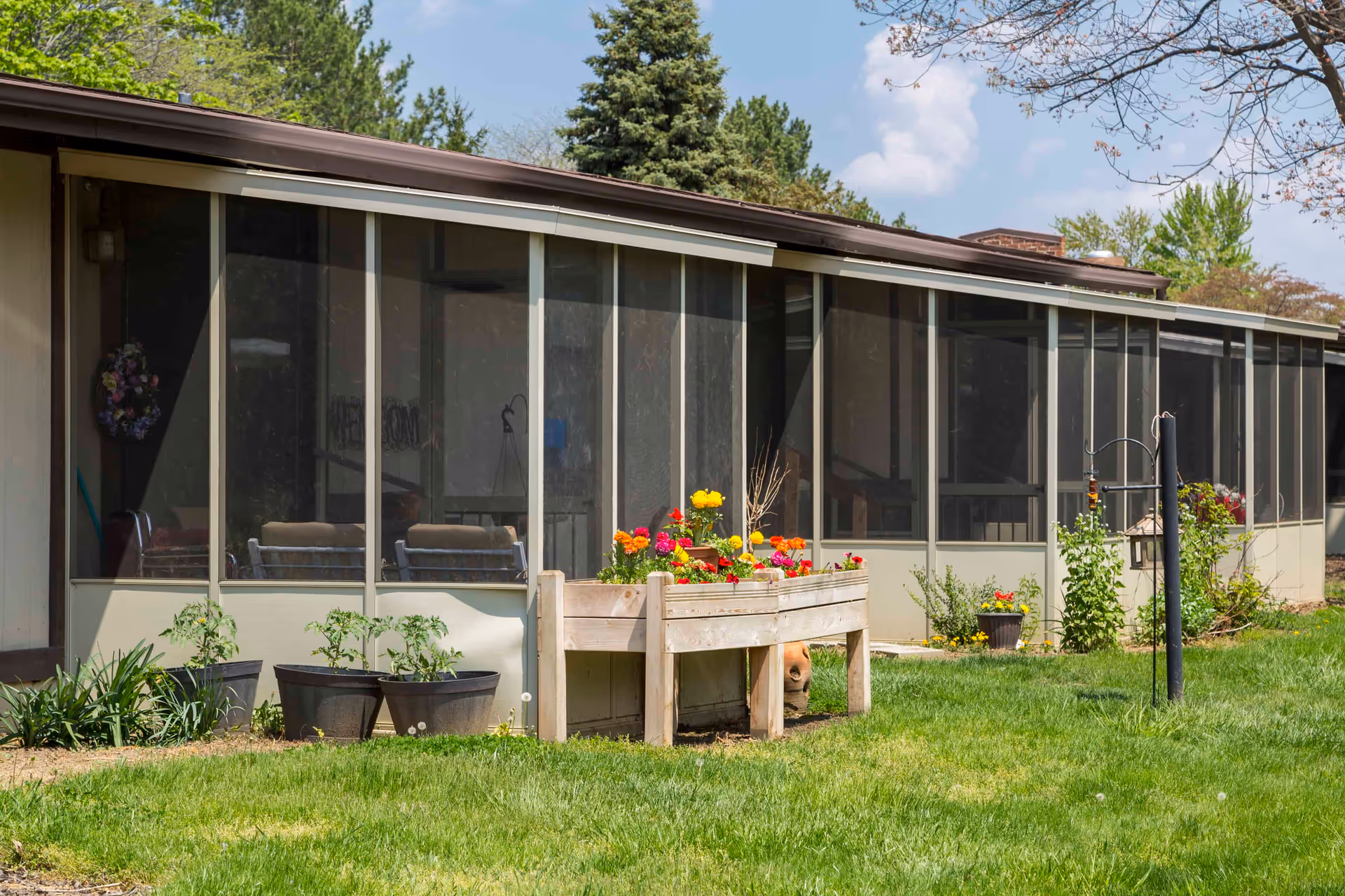 A screened-in porch attached to a building with several potted plants and a raised wooden planter box filled with colorful flowers in front. The porch has multiple sections with mesh screens and cushioned chairs visible inside. The surrounding area has green grass, some shrubs, and trees under a partly cloudy sky.