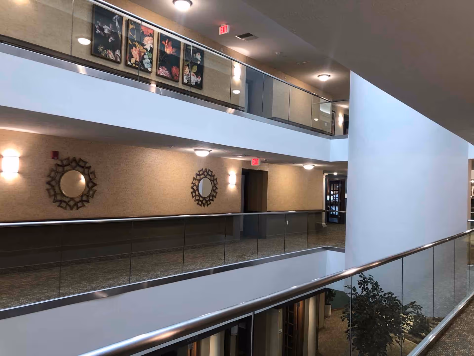 Interior view of a multi-level hallway in a retirement community with glass railings, decorative mirrors on the walls, framed floral artwork, ceiling lights, and exit signs.