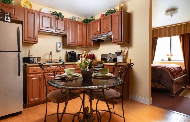 A cozy kitchen and dining area with wooden cabinets, a stainless steel refrigerator, a round table set for two with green bowls and plates, and a floral centerpiece. In the background, a bedroom with a bed, window, and curtains is partially visible.