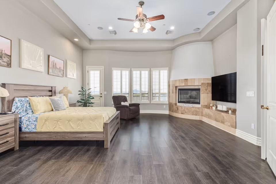 A spacious bedroom with a wooden bed frame and yellow bedding, a nightstand with a lamp, a reclining chair near large windows with white shutters, a curved fireplace with beige tiles, and a mounted flat-screen TV on the wall. The room has dark wood flooring and a ceiling fan with lights.