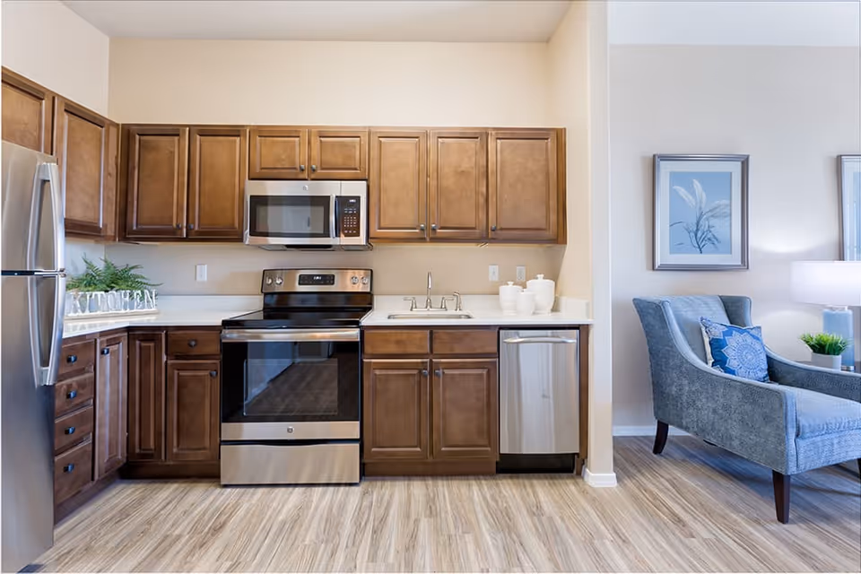 A modern kitchen with wooden cabinets, stainless steel appliances including a refrigerator, oven, microwave, and dishwasher. The countertop has a small plant and decorative jars. To the right, part of a living area is visible with a blue upholstered armchair, a side table with a lamp, and framed artwork on the wall.