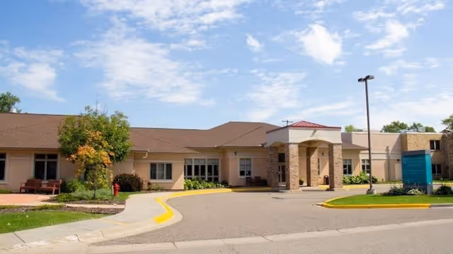 Single-story senior living facility building front with a covered entrance driveway, manicured landscaping, and a clear blue sky.